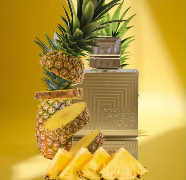 Złoty, prostokątny flakon perfum Haramain Amber Oud Extreme stojący na żółtym tle obok całego ananasa, pokrojonych plastrów i kawałków tego owocu.