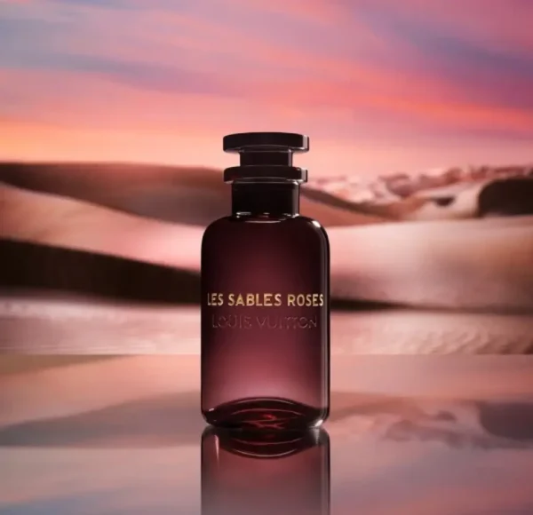 Flakon perfum Louis Vuitton Les Sables Roses w odcieniu głębokiego burgundu, stojący na lustrzanej powierzchni, która odbija pustynny krajobraz. W tle piaszczyste wydmy o zachodzie słońca z różowo-pomarańczowym niebem.