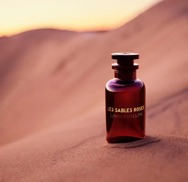 Butelka perfum Louis Vuitton Les Sables Roses umieszczona na piaszczystej wydmie. Światło zachodzącego słońca oświetla flakon i piaszczyste tło.