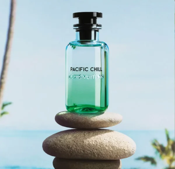 Flakon perfum Louis Vuitton Pacific Chill ustawiony na stosie gładkich, jasnych kamieni. W tle błękitne niebo i zarys plaży z palmami.