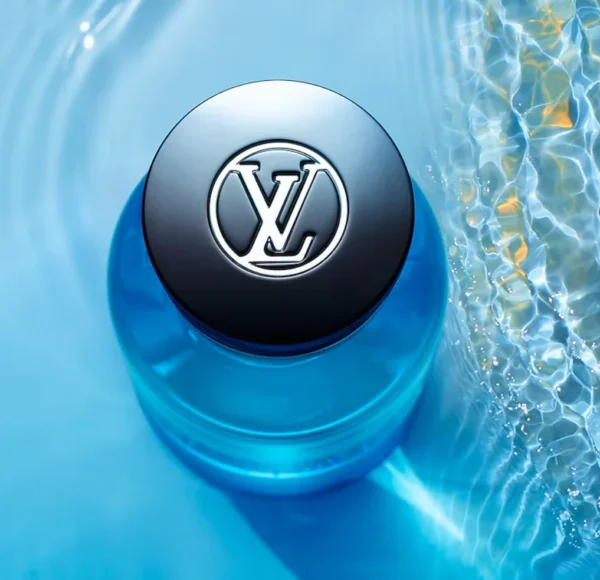 Ujęcie z góry na czarny korek flakonu perfum Louis Vuitton Afternoon Swim z wyraźnym srebrnym logo LV, otoczony błękitną wodą z falami i odbiciami światła.