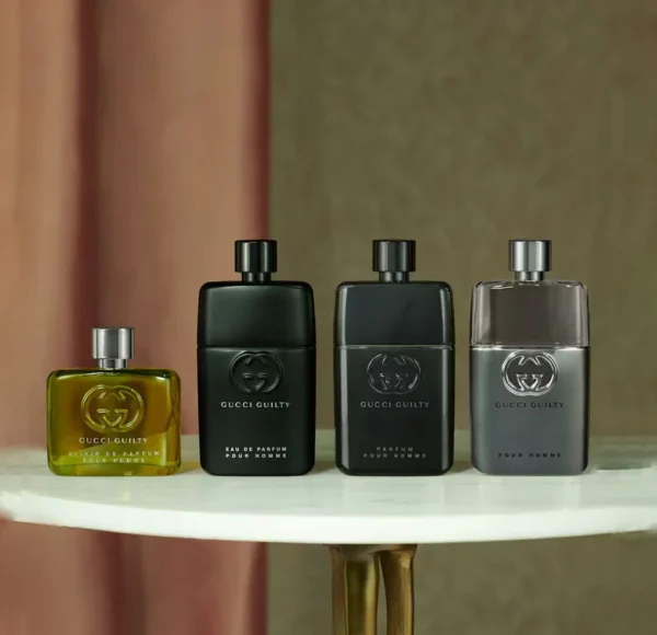 Cztery flakony perfum Gucci Guilty Pour Homme (zielony Elixir, czarne Eau de Parfum i Parfum, oraz szary Pour Homme) ustawione w rzędzie na jasnym, okrągłym stole.