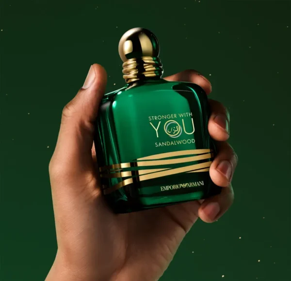 Dłoń trzymająca flakon perfum Emporio Armani Stronger with You Sandalwood, w kolorze ciemnozielonym ze złotymi napisami i korkiem, na rozmytym, zielonym tle.