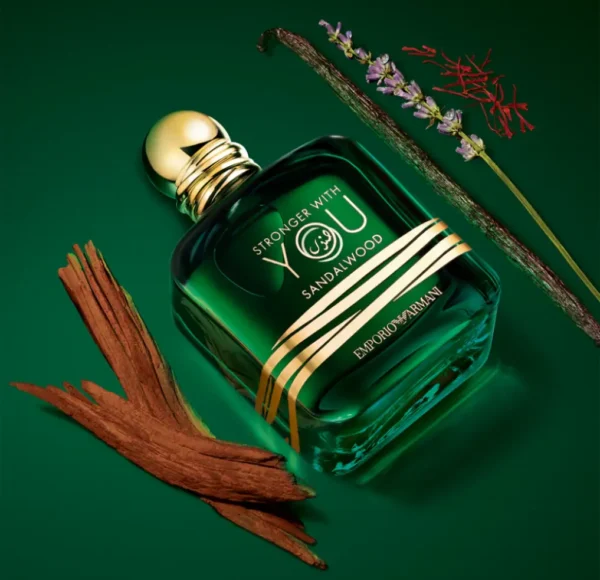 Flakon perfum Emporio Armani Stronger with You Sandalwood, z kawałkiem drewna sandałowego, laską wanilii i suszonymi kwiatami na zielonym podłożu.